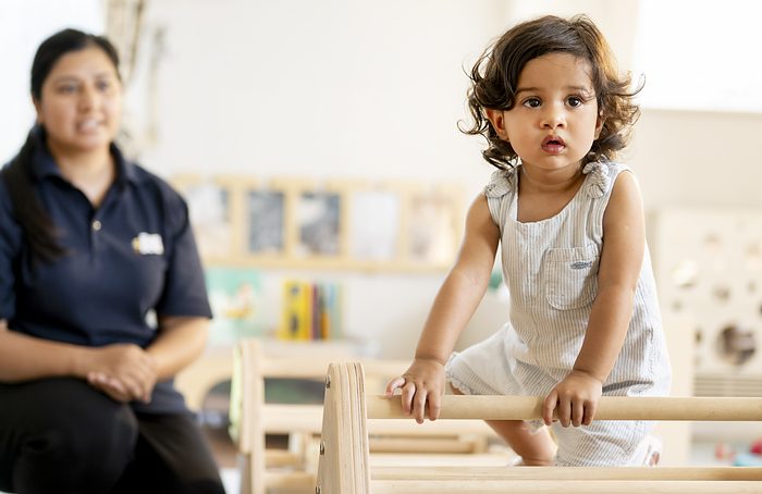 A practitioner supervising a young child climbing on low wooden play equipment.